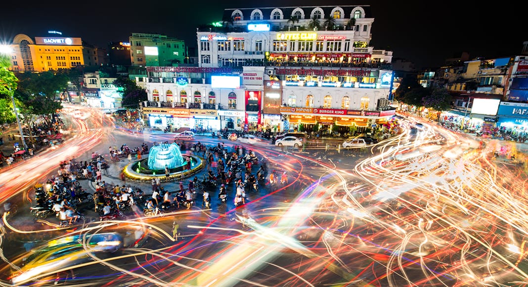 People and vehicles light trails crowd on busy intersection locating next to Hoan Kiem lake.